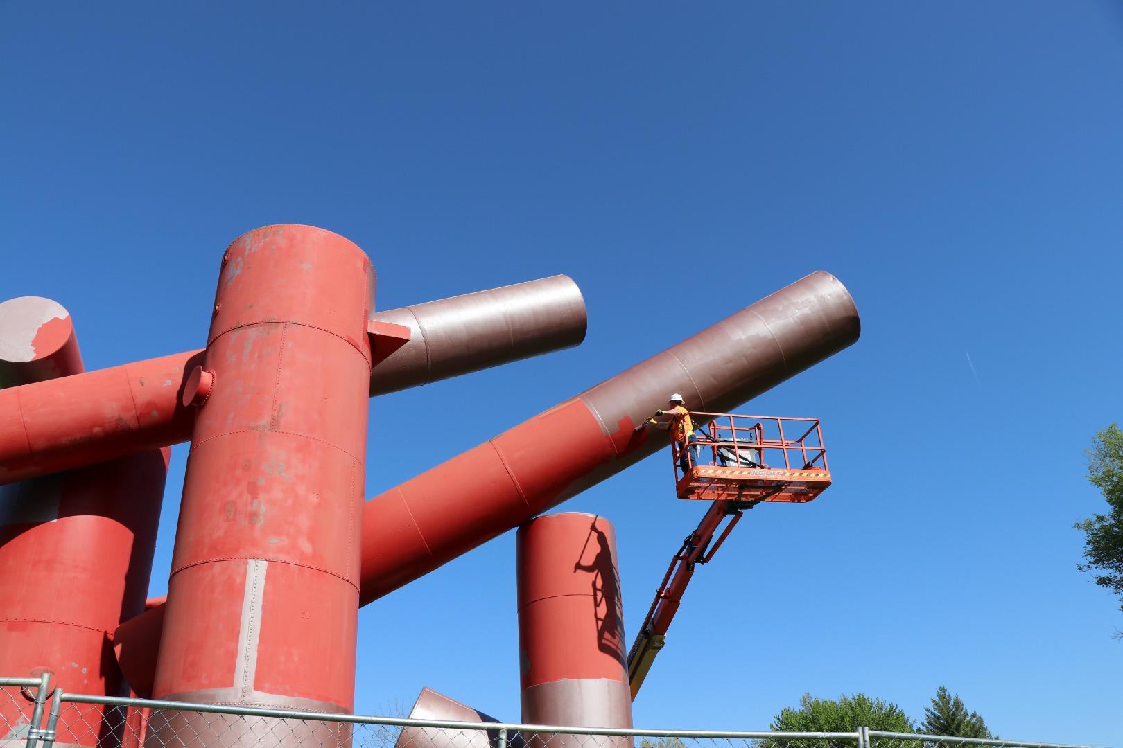 A painter in a man lift applies a silver primer to the surface of a large sculpture made of reclaimed steel tanks.